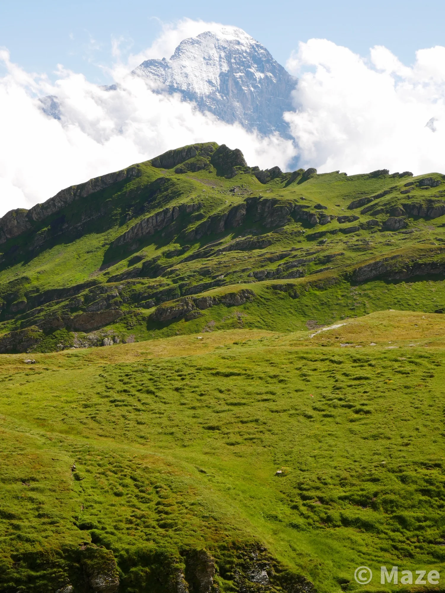 A mountain landscape image from Grindelwald, Switzerland. The foreground is dominated by lush, rolling green grassy hills, and the background is pierced by the white and blues of a distant mountain draped in clouds against a bright blue sky.&nbsp;