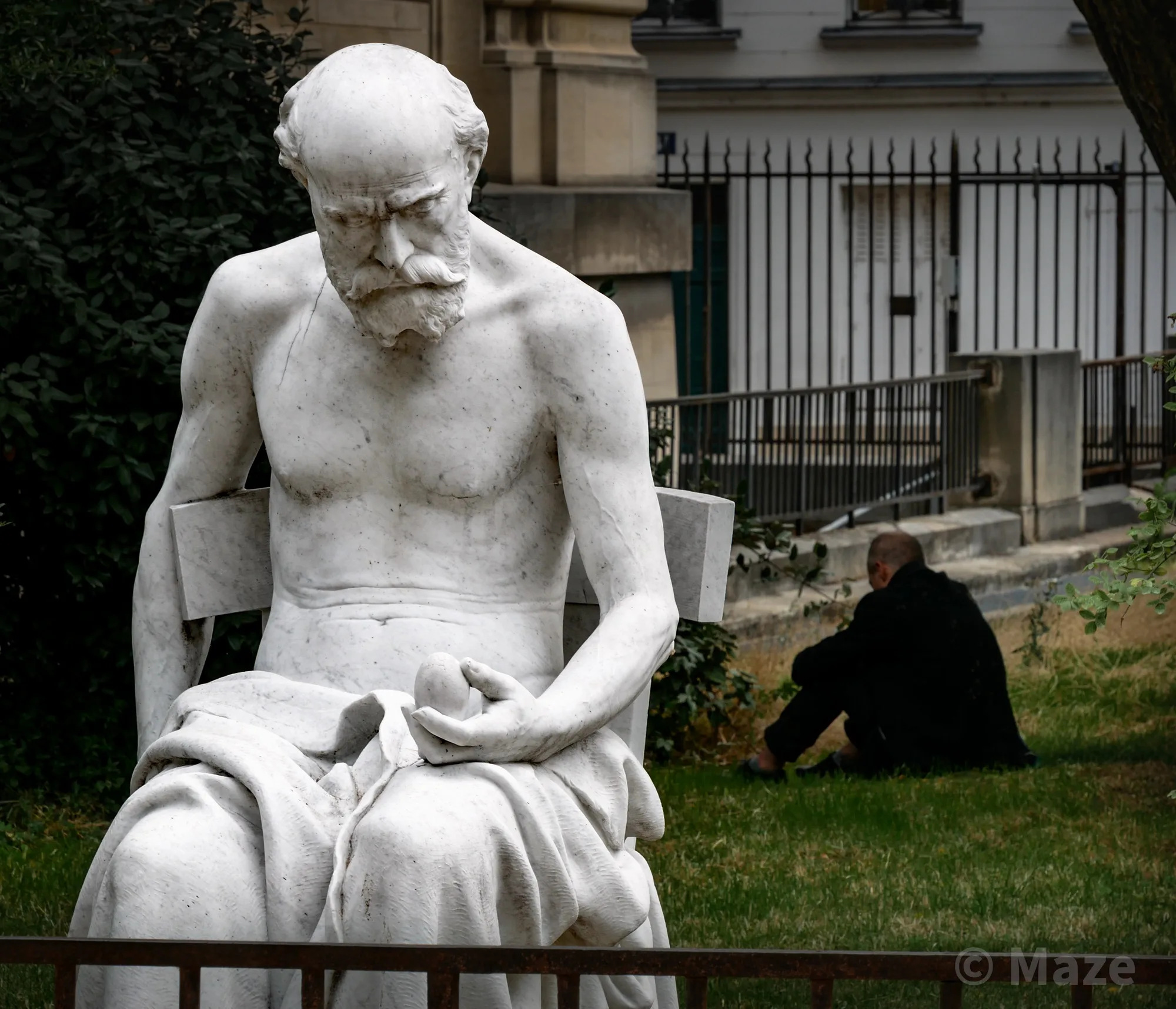 A man in deep contemplation sitting behind the Science et Mystère sculpture in the Jardin des Plantes in Paris, France (July 2025).